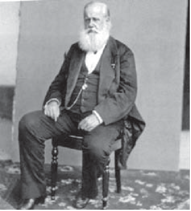 Fotografia em preto e branco: D. Pedro II, idoso, de barba branca longa, sentado em uma cadeira, com terno escuro e colete, as mãos apoiadas nas pernas, diante de um painel liso.