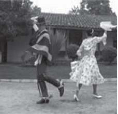Fotografia em preto e branco de um casal dançando cueca ao ar livre, diante de uma casa de telhado de telhas: o homem, de chapéu, poncho e botas, e a mulher, de vestido rodado estampado, ambos com um braço erguido agitando um lenço.
