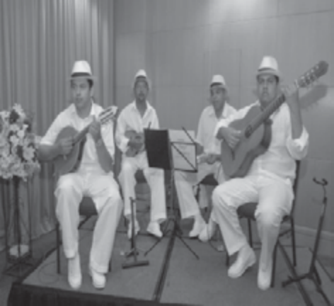 Fotografia de três músicos de chapéu e roupas brancas, sentados, tocando cavaquinho, pandeiro e violão.