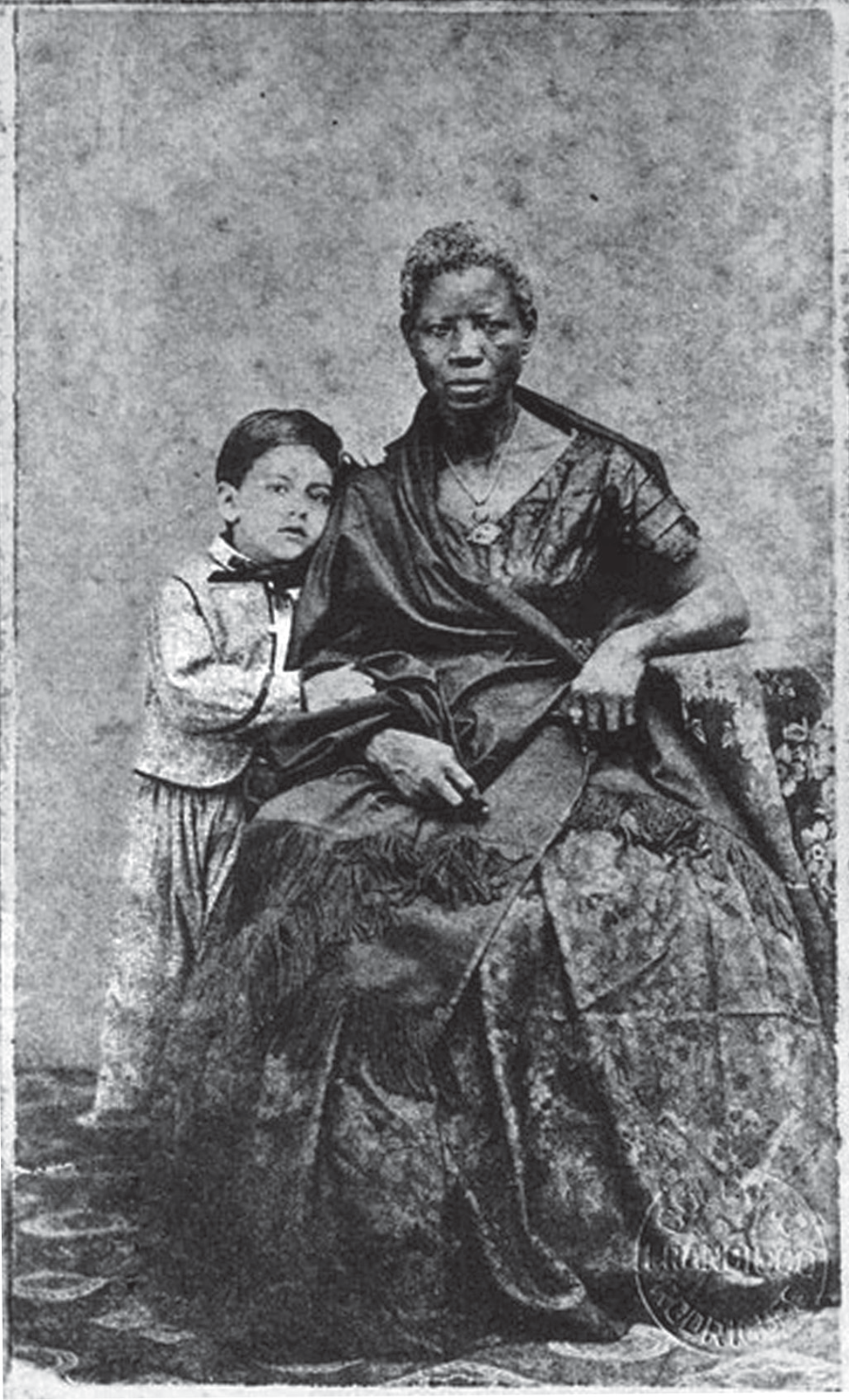 Fotografia de estúdio em preto e branco, do século XIX, com bordas desgastadas. Uma mulher negra, de expressão séria, está sentada, vestida com um vestido escuro de mangas compridas, xale com franjas e um cordão com pingente ao pescoço. Ao lado dela, em pé, um menino branco de cabelos escuros, com casaco claro, gravata-borboleta e calças, apoia o braço no colo dela. Fundo neutro de estúdio; no canto inferior direito, carimbo do fotógrafo.