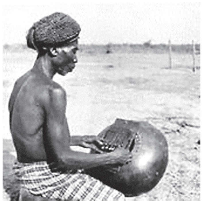 Fotografia em preto e branco de um homem sentado ao ar livre, com o tronco nu e uma faixa na cabeça, tocando com os polegares um instrumento de lâminas metálicas montado sobre uma grande cabaça arredondada apoiada no colo; ao fundo, um terreno aberto e plano.