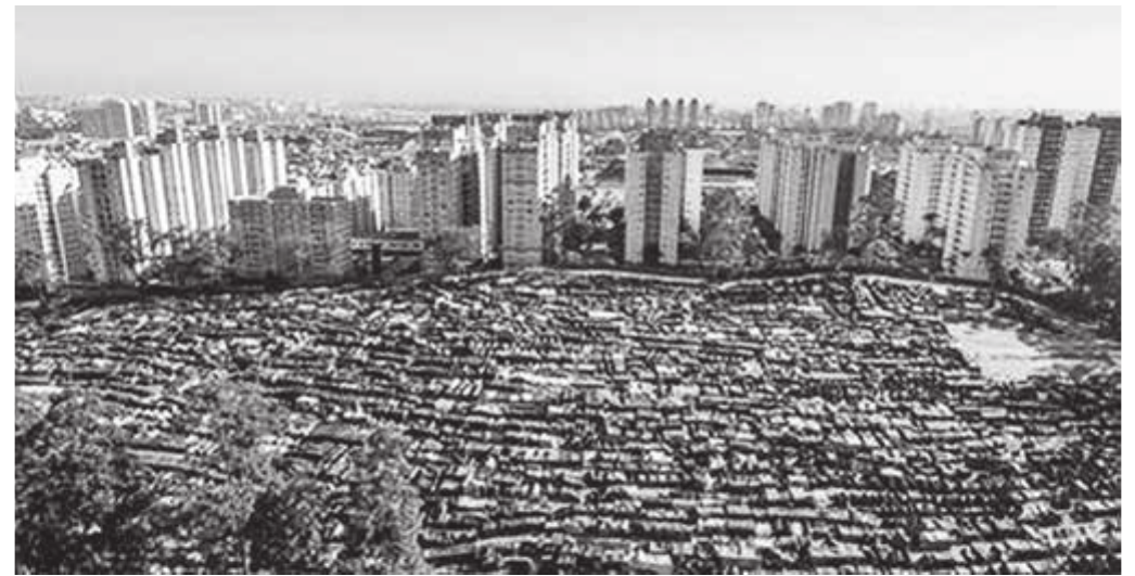 Fotografia aérea em preto e branco: em primeiro plano, uma extensa área coberta por milhares de barracas de lona, muito próximas umas das outras, com algumas árvores e um trecho de terreno vazio; ao fundo, separada por uma faixa de vegetação, uma sequência de edifícios residenciais altos e, no horizonte, a mancha urbana da cidade.
