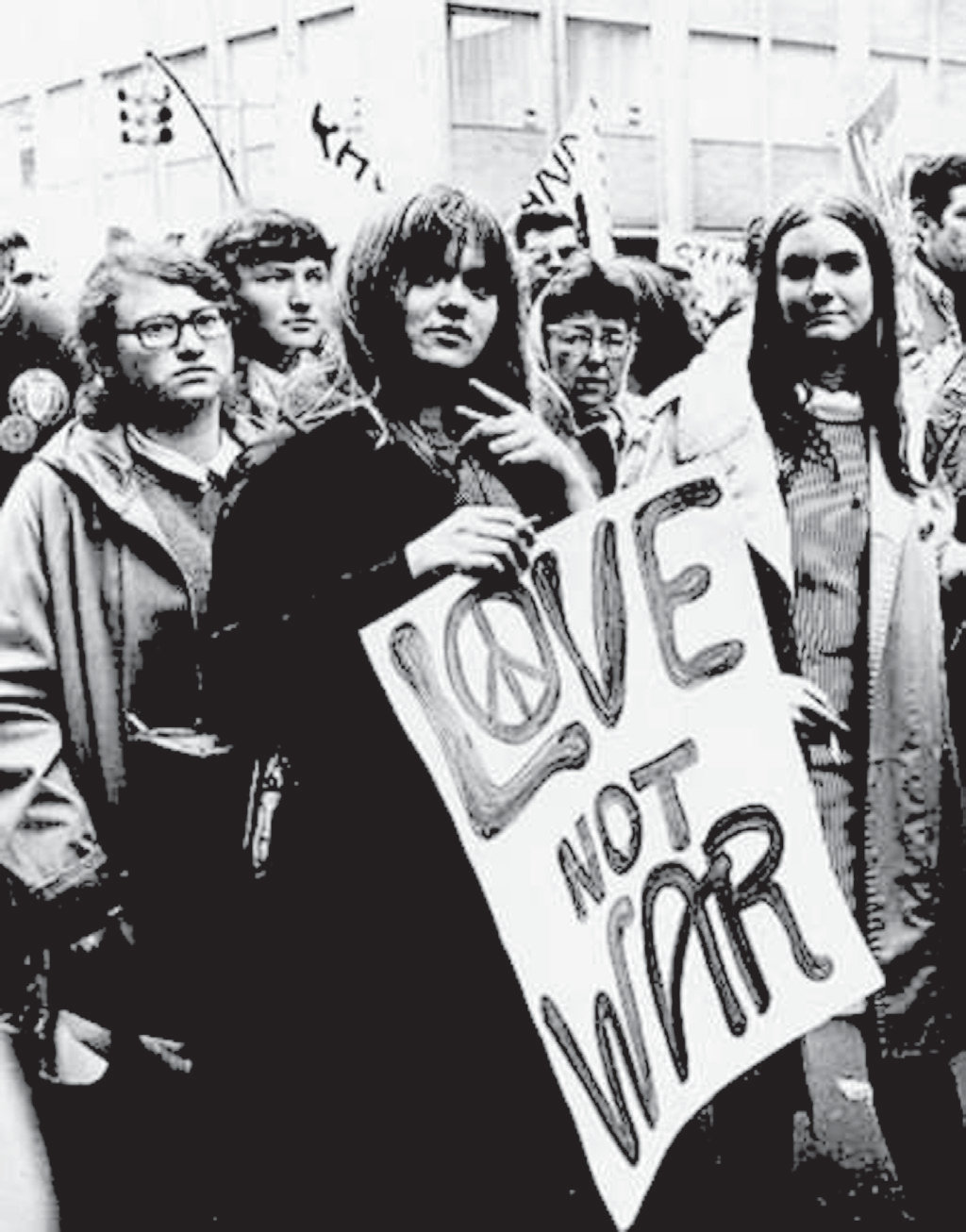 Fotografia em preto e branco de uma manifestação de rua: um grupo de jovens, alguns de óculos e cabelos compridos, caminha em direção à câmera; uma moça à frente segura um cartaz escrito LOVE NOT WAR, com o símbolo da paz desenhado dentro da letra O.