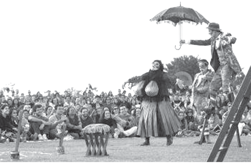 Fotografia em preto e branco de um espetáculo ao ar livre: atores em figurinos de época (uma atriz de saia longa e blusa escura, de braços abertos, e um ator de chapéu e casaco florido, sobre pernas de pau, segurando um guarda-chuva) atuam sobre a grama, com um banquinho e uma escada como cenário; ao fundo, uma grande plateia sentada no gramado e árvores.