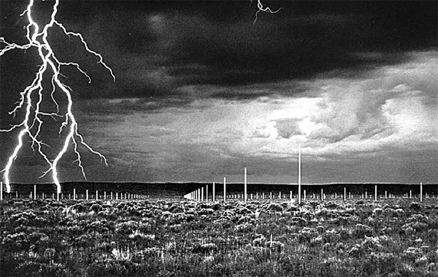 Fotografia em preto e branco de um campo plano com vegetação rasteira, sob céu de tempestade. Dezenas de hastes metálicas finas e verticais, dispostas em fileiras regulares, cobrem o campo; à esquerda, raios caem do céu escuro.