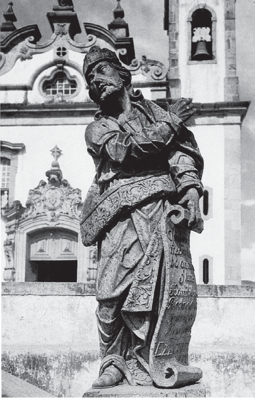 Fotografia em preto e branco de uma escultura em pedra-sabão de Aleijadinho: um profeta com turbante e manto com muitas dobras, segurando um pergaminho com inscrição, sobre o muro do adro; ao fundo, a fachada da igreja do Santuário do Bom Jesus de Matosinhos, em Congonhas.