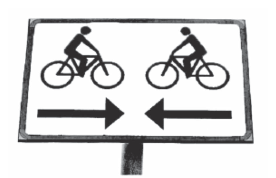 Fotografia em preto e branco de uma placa de trânsito retangular, de fundo branco e moldura preta, fixada em um poste. Na placa há dois pictogramas iguais de um ciclista pedalando, lado a lado, ambos voltados para a direita. Abaixo de cada ciclista há uma seta larga: a seta da esquerda aponta para a direita e a seta da direita aponta para a esquerda, uma de frente para a outra.
