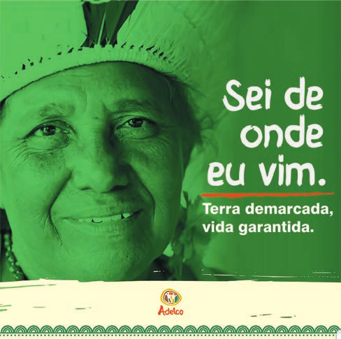 Cartaz de campanha em tons de verde. Ao fundo, o retrato sorridente de uma mulher indígena idosa, com adorno de penas na cabeça e colar. À direita, em letras brancas grandes: “Sei de onde eu vim.” e, abaixo, em destaque: “Terra demarcada, vida garantida.”. No rodapé, sobre faixa clara, a marca “Adelco” e uma barra decorativa com grafismo indígena.