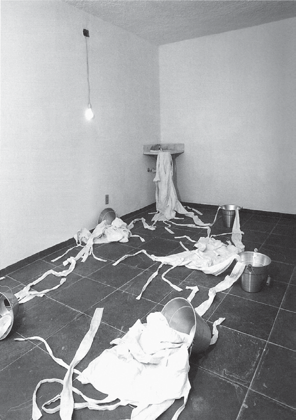 Fotografia de uma sala com paredes brancas e piso escuro de lajotas: uma lâmpada nua pendurada no alto à esquerda, uma pia de canto com uma camisa de força pendurada, baldes de metal espalhados e várias camisas de força brancas com tiras longas estendidas pelo chão, como se escorressem dos baldes.