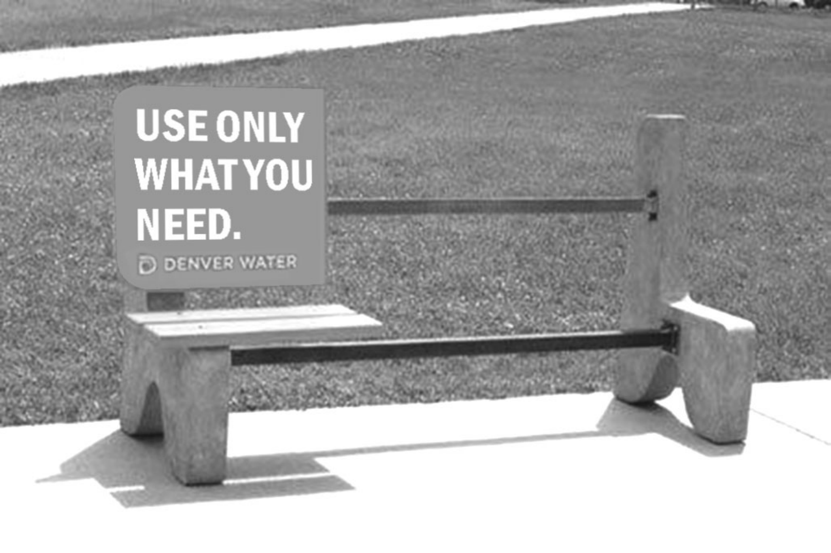 Fotografia de um banco de concreto em um parque: as barras metálicas atravessam o banco, mas só um terço do assento tem ripas de madeira; no encosto, uma placa cinza com o texto "USE ONLY WHAT YOU NEED." e o logotipo "DENVER WATER".