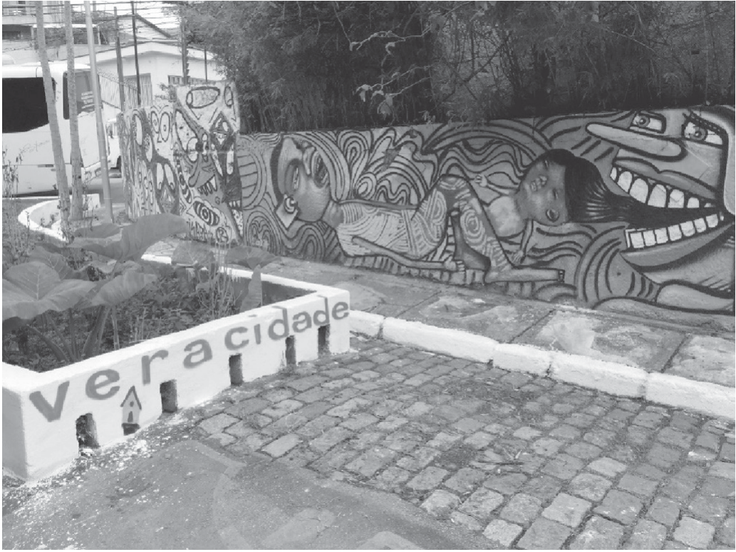 Fotografia em preto e branco de uma viela calçada com paralelepípedos. Ao fundo, um muro comprido inteiramente coberto por grafites, com figuras humanas estilizadas, rostos e uma grande boca aberta com dentes à mostra. Em primeiro plano, a mureta branca de um canteiro com plantas traz pintada a inscrição “vera cidade” e pequenos desenhos de casas. À esquerda, parte de um veículo estacionado e, acima do muro, árvores.