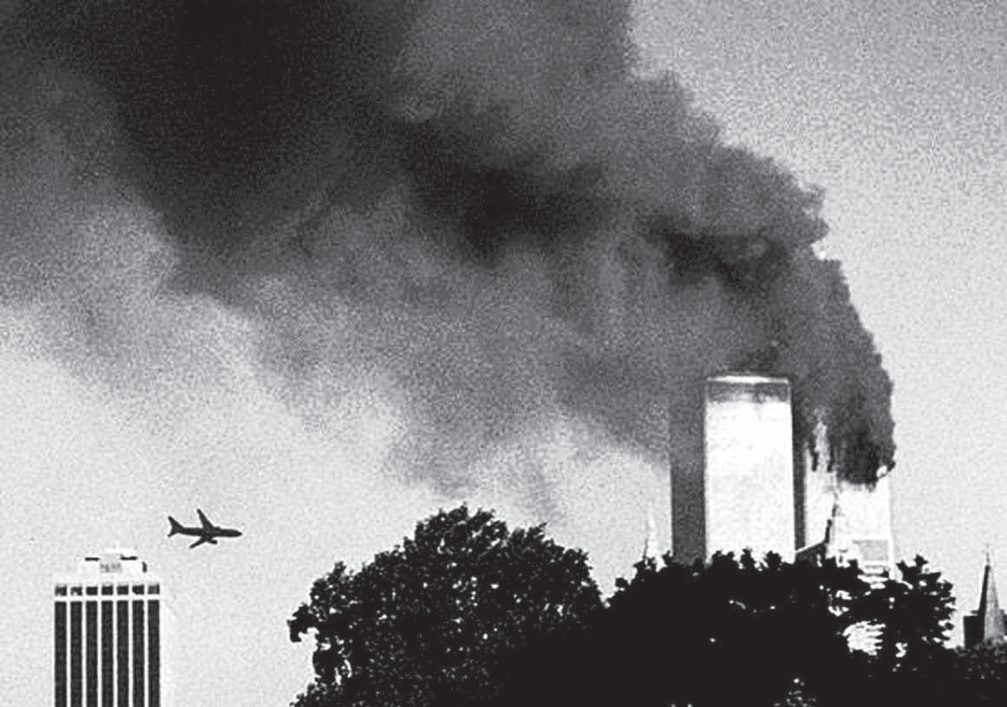Fotografia em preto e branco de Nova York em 11 de setembro de 2001: as duas torres do World Trade Center ao fundo, uma delas com uma enorme coluna de fumaça escura que se espalha pelo céu; à esquerda, um avião de passageiros aparece voando em direção às torres; no primeiro plano, copas de árvores e outros edifícios da cidade.