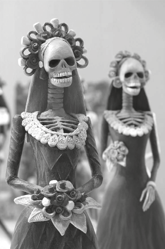 Fotografia de duas estatuetas de esqueletos vestidos como mulheres, no estilo das catrinas mexicanas: a da frente, em foco, usa vestido longo, gola rendada, coroa de flores e segura um buquê; a de trás, desfocada, também usa vestido e coroa de flores.