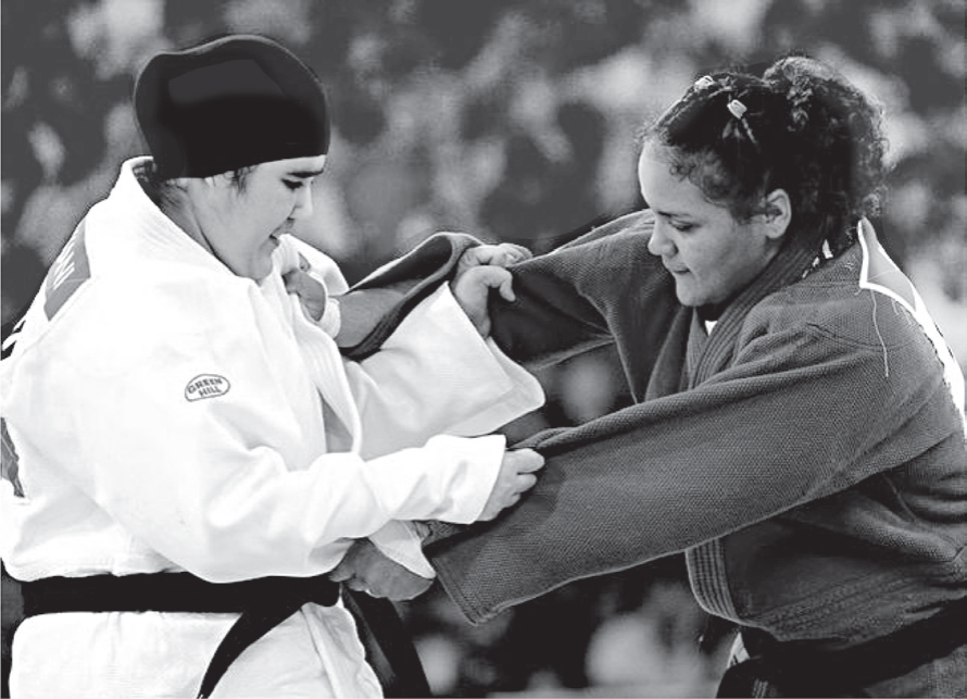 Fotografia em preto e branco de uma luta de judô. À esquerda, uma judoca de quimono branco e faixa preta usa um véu escuro que cobre os cabelos; à direita, uma adversária de quimono escuro e cabelos presos. As duas seguram a gola e a manga uma da outra, diante da plateia desfocada.