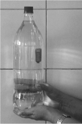 Fotografia da mesma garrafa PET sendo apertada pela mão; o pequeno frasco escuro aparece agora na metade inferior da garrafa, deslocado para baixo.