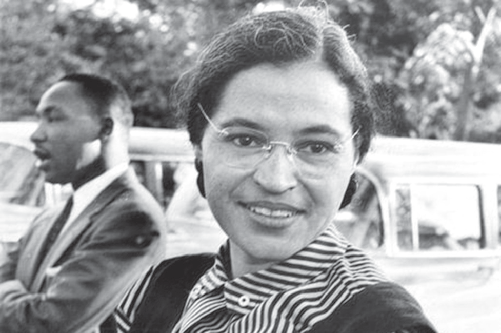 Fotografia em preto e branco: em primeiro plano, Rosa Parks, mulher negra de óculos, cabelo preso e blusa listrada, sorri para a câmera; atrás dela, à esquerda, Martin Luther King Jr., de terno, olha para o lado; ao fundo, automóveis antigos e árvores.