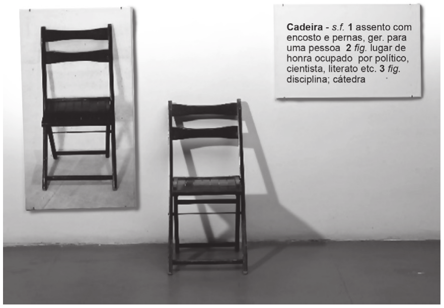 Fotografia da instalação numa sala de paredes claras: à esquerda, presa à parede, a fotografia ampliada de uma cadeira dobrável de madeira; ao centro, no chão, a mesma cadeira dobrável; à direita, na parede, um painel branco com a definição de dicionário do verbete.

Cadeira - s.f. 1 assento com encosto e pernas, ger. para uma pessoa 2 fig. lugar de honra ocupado por político, cientista, literato etc. 3 fig. disciplina; cátedra
