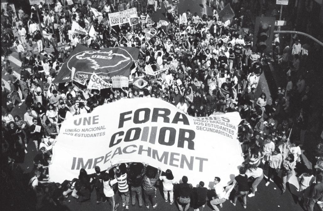 Fotografia aérea em preto e branco de uma passeata que ocupa uma avenida. No centro, jovens seguram uma grande faixa branca com os dizeres 'UNE – União Nacional dos Estudantes', 'UBES – União Brasileira dos Estudantes Secundaristas', 'FORA COLLOR' em letras grandes e 'IMPEACHMENT' abaixo. Ao redor, uma multidão com cartazes, faixas menores, uma bandeira da UBES com o mapa do Brasil e uma bandeira do Brasil.