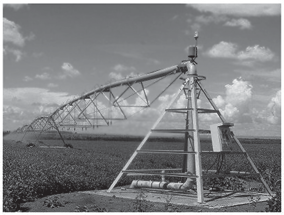Fotografia de um pivô central de irrigação num campo: a torre central com a tubulação vertical e o longo cano horizontal apoiado em torres com rodas, sob céu com nuvens.