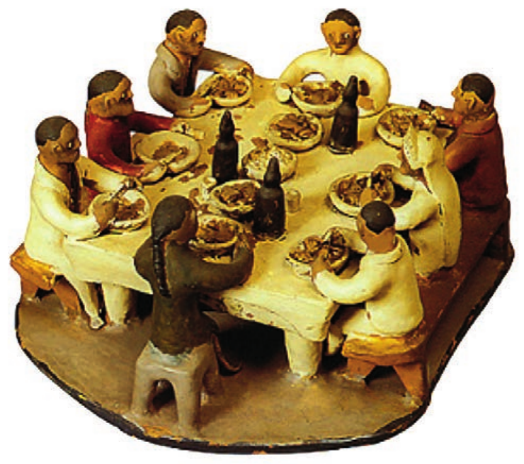 Fotografia de uma escultura em cerâmica policromada: sobre uma base irregular de barro, uma mesa retangular clara com sete pessoas sentadas ao redor, em bancos, cada uma diante de um prato com comida; sobre a mesa há garrafas escuras e canecas. As figuras vestem camisas brancas ou vermelhas e calças escuras; uma delas, de costas para o observador, está sentada em um banquinho cinza.