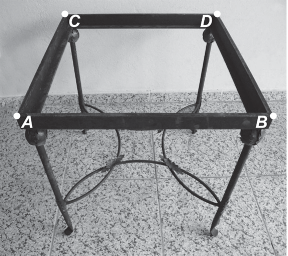 Fotografia de uma base de mesa de ferro, retangular, com quatro pés; os vértices do retângulo superior estão marcados com as letras A (frente esquerda), B (frente direita), C (fundo esquerda) e D (fundo direita).