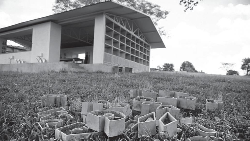 Fotografia em preto e branco: em primeiro plano, sobre a grama, vasos de cerâmica em forma de letras, com terra e plantas brotando, dispostos de modo a compor palavras; ao fundo, um galpão branco com telhado inclinado e uma grande estante de nichos na fachada, onde ficam as letras-vaso.