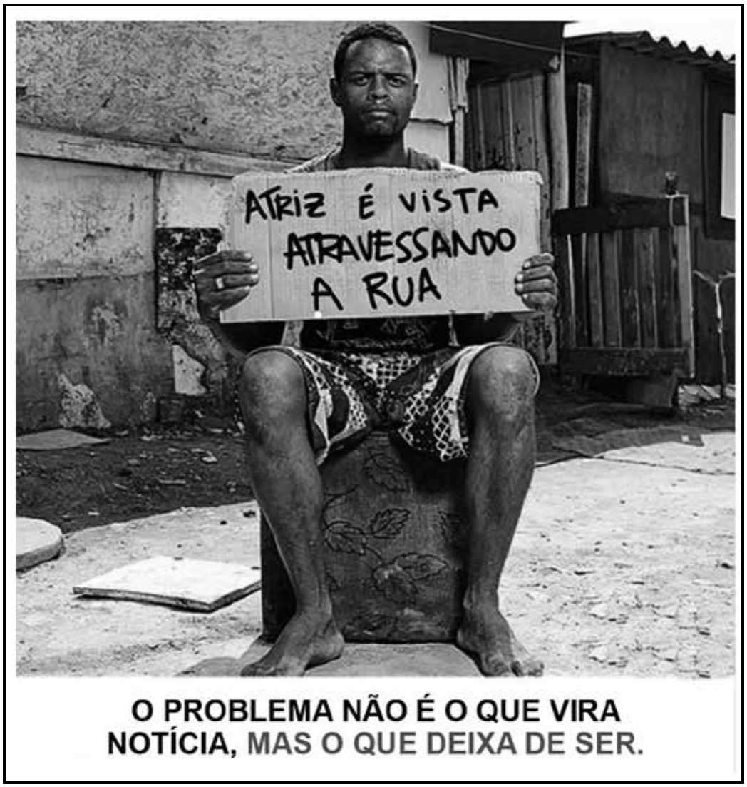 Anúncio em preto e branco, com moldura. Fotografia de um homem negro, descalço e de bermuda, sentado sobre um caixote em uma rua de terra, diante de barracos de madeira; ele olha para a câmera e segura um pedaço de papelão em que se lê, escrito à mão: “ATRIZ É VISTA ATRAVESSANDO A RUA”. Na faixa inferior, o texto: “O PROBLEMA NÃO É O QUE VIRA NOTÍCIA, MAS O QUE DEIXA DE SER.”, com a segunda parte em cinza.