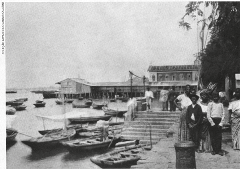 Fotografia antiga em preto e branco de um cais em Salvador: à esquerda, vários barcos a remo e a vela atracados junto a uma escadaria de pedra; ao fundo, um galpão sobre estacas e um sobrado; à direita, sobre o cais, um grupo de pessoas, homens e mulheres negros e mestiços, algumas mulheres com saias longas e turbantes, ao lado de um poste de amarração. Na margem esquerda, a inscrição vertical “Coleção Apparecido Jannir Salatini”.