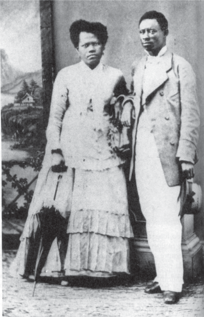 Fotografia de estúdio, em preto e branco, de um casal negro em pé diante de um cenário pintado com paisagem: a mulher usa um vestido longo e claro com babados e segura uma sombrinha fechada; o homem usa paletó longo claro, calça clara e segura um chapéu na mão; ambos estão calçados.