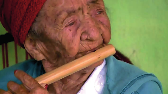 Fotografia em close de uma senhora idosa, de lenço vermelho na cabeça e blusa azul-clara, tocando um pífano de madeira clara que segura junto à boca (vê-se uma das mãos); ao fundo, parede verde.