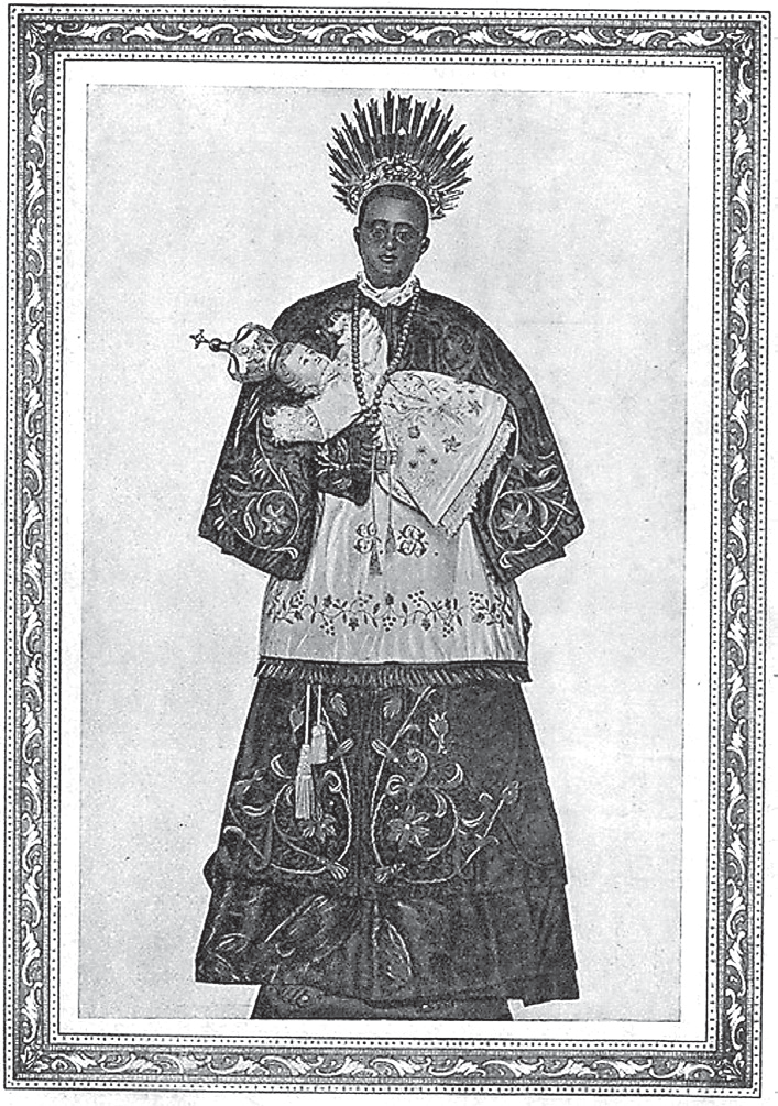 Fotografia antiga, emoldurada, de uma imagem de São Benedito: um santo negro, com resplendor de raios na cabeça, vestido com hábito e manto ricamente bordados, segurando o Menino Jesus no braço esquerdo.