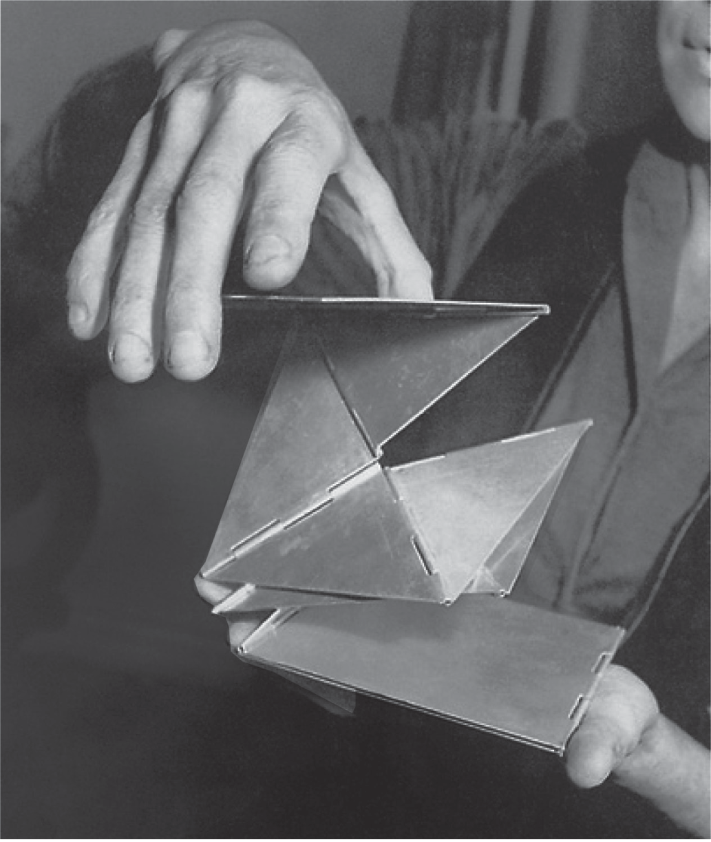 Fotografia em preto e branco. Duas mãos seguram e dobram um pequeno objeto formado por placas triangulares de metal unidas por dobradiças, que se abrem em várias direções como um origami metálico.