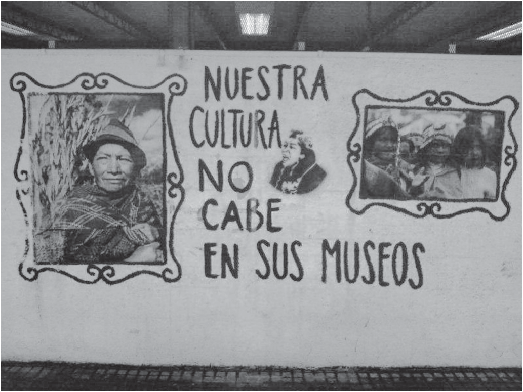 Fotografia em preto e branco de um muro branco pintado, sob uma cobertura. No centro, em letras grandes desenhadas à mão, a frase em espanhol: “NUESTRA CULTURA NO CABE EN SUS MUSEOS”, com um pequeno rosto de mulher pintado ao lado da palavra “CULTURA”. À esquerda e à direita, dentro de molduras ornamentadas pintadas no muro, há dois retratos: no da esquerda, uma pessoa indígena de chapéu e manta, diante de uma plantação; no da direita, um grupo de pessoas indígenas com gorros e roupas tradicionais.