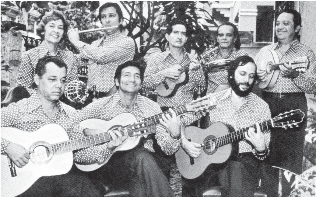 Fotografia em preto e branco de um grupo de choro com oito músicos, todos de camisa estampada. Na fila da frente, três homens sentados tocam violões. Atrás, da esquerda para a direita: uma mulher segura um pandeiro erguido; um homem toca uma flauta transversal; um homem toca um cavaquinho; um homem segura um bandolim; e, à direita, um homem segura outro instrumento de percussão de mão. Ao fundo, plantas e uma parede.