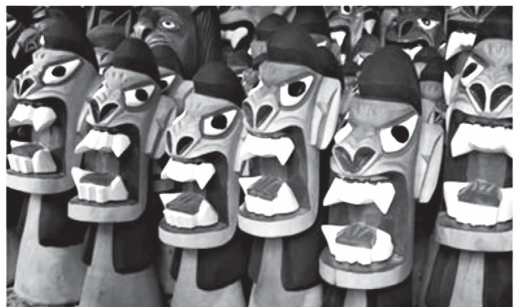 Fotografia em preto e branco de várias carrancas de madeira enfileiradas, esculturas de rostos fantásticos com olhos arregalados, narizes grandes, bocas abertas mostrando dentes pontudos e cabelos esculpidos; as figuras têm pinturas em faixas e estão colocadas uma ao lado da outra, em várias fileiras.