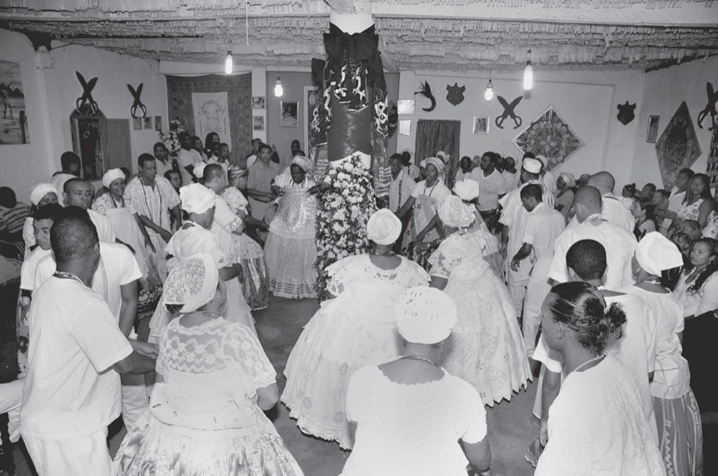 Fotografia em preto e branco de um salão de cerimônia com bandeirinhas de papel no teto, lâmpadas penduradas e objetos decorativos nas paredes; dezenas de pessoas vestidas de branco — mulheres com saias rodadas de renda e panos amarrados na cabeça, homens de camisa e calça brancas — dançam em roda ao redor de um pilar central envolto em tecidos e enfeitado com flores.