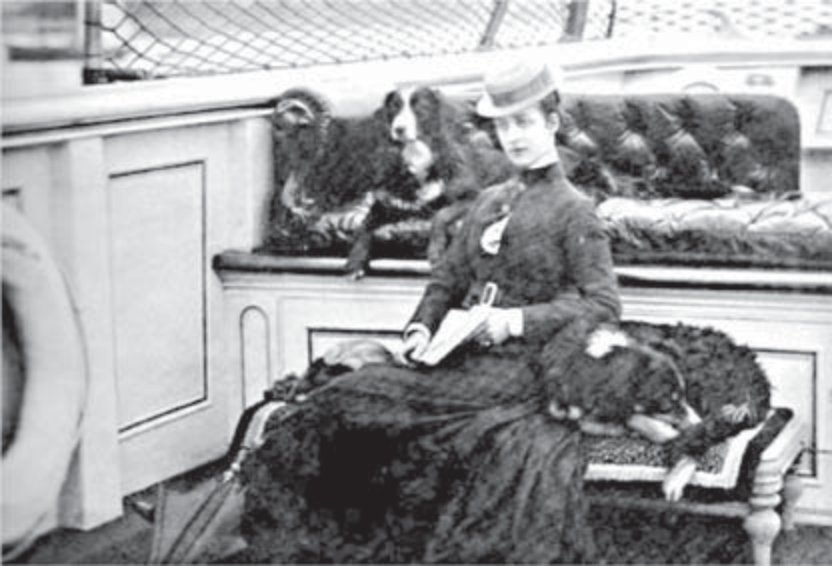 Fotografia em preto e branco da princesa Alexandra, do século XIX: mulher sentada em um banco de jardim, com vestido longo e escuro que cobre todo o corpo, chapéu, um pequeno livro nas mãos e dois cães ao seu lado; postura rígida e formal.