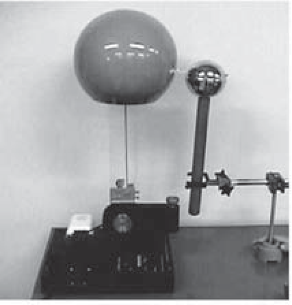 Fotografia de um gerador de Van de Graaff: uma esfera metálica grande sobre uma coluna, ao lado de uma esfera menor montada em uma haste, em uma bancada de laboratório.