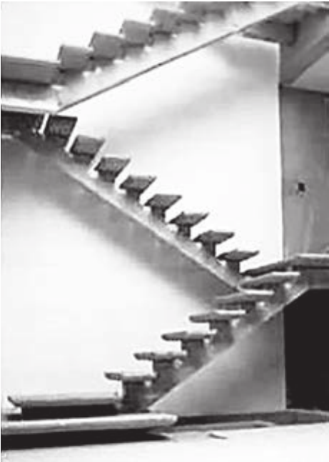 Fotografia em preto e branco de uma escada de concreto em construção, com degraus em balanço projetados de uma parede lisa e uma viga inclinada de apoio, vista de lado num ambiente interno.