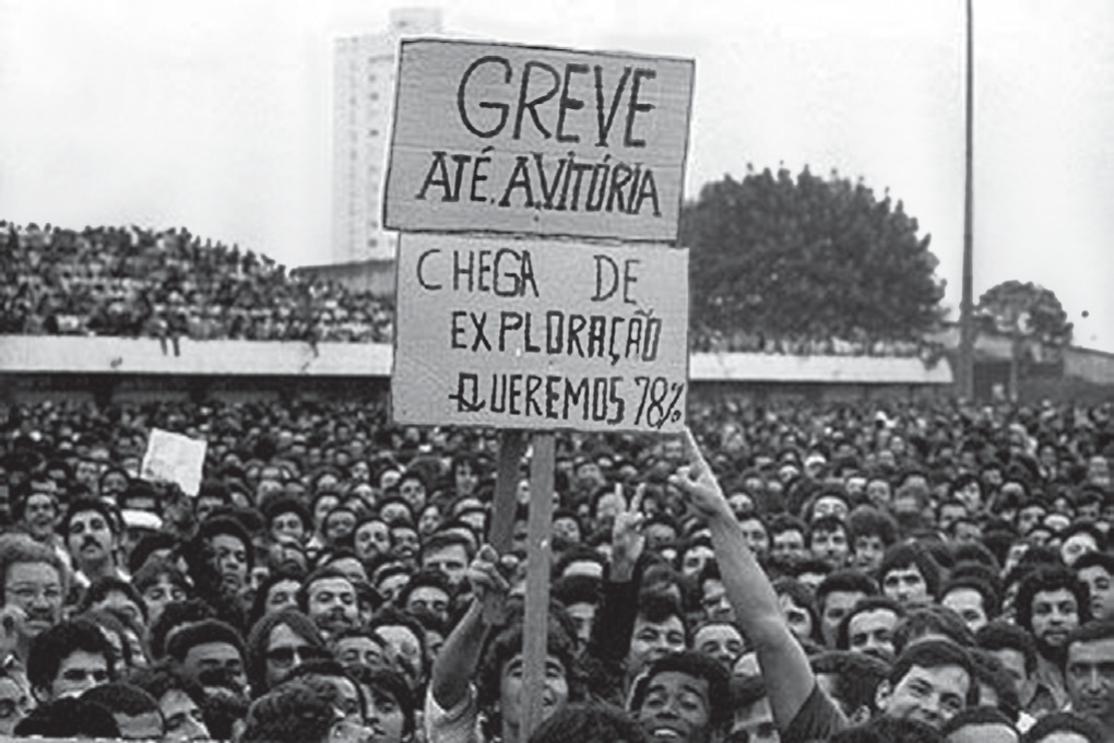 Fotografia em preto e branco de uma multidão de trabalhadores reunida em um estádio; ao centro, um cartaz erguido com os dizeres 'Greve até a vitória' e 'Chega de exploração, queremos 78%'.