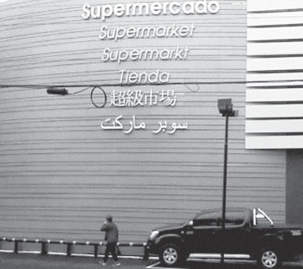 Fotografia em preto e branco da fachada de um supermercado: na parede alta, uma abaixo da outra, as inscrições “Supermercado”, “Supermarket”, “Supermarkt”, “Tienda”, “超級市場” (em caracteres chineses) e “سوبر ماركت” (em escrita árabe). Na calçada, uma pessoa caminha junto a uma grade, um poste de iluminação e uma caminhonete estacionada.