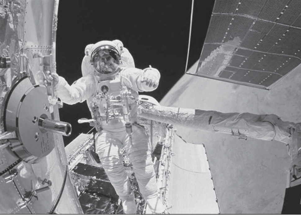 Fotografia em preto e branco de um astronauta em traje espacial trabalhando fora de uma nave, próximo a uma estrutura cilíndrica com uma escotilha aberta e a um painel solar, com o espaço escuro ao fundo.