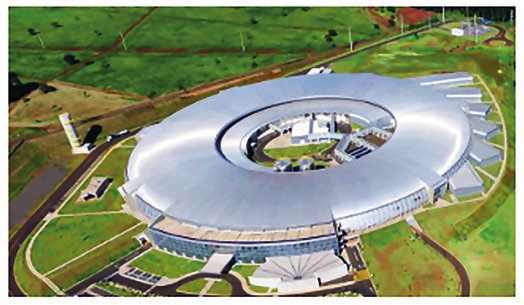 Fotografia aérea de um acelerador de partículas: um edifício circular de cobertura metálica prateada, em forma de anel, cercado por gramado, estacionamento e vias de acesso, com vegetação e construções menores ao redor.