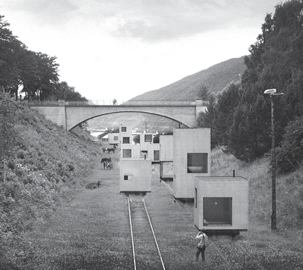 Fotografia em preto e branco de um vale com montanhas: casas em forma de caixa, com janelas, apoiadas sobre trilhos de trem que atravessam a vegetação; ao fundo, uma ponte de concreto em arco; uma pessoa caminha ao lado das casas.