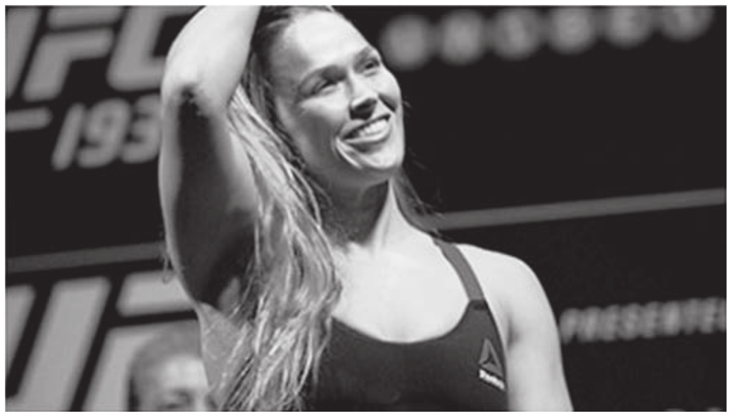 Fotografia em preto e branco de uma atleta loira sorrindo, de top esportivo, com um braço erguido sobre a cabeça, diante de um painel de evento de artes marciais.