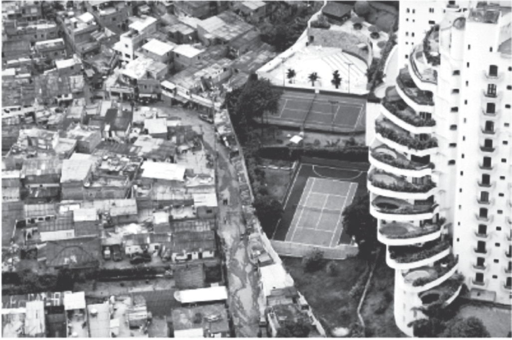 Fotografia aérea em preto e branco. À esquerda, uma favela densa, com centenas de casas pequenas de telhados irregulares e ruas estreitas. À direita, separado por um muro, um conjunto residencial de luxo: um prédio alto e curvo com varandas ajardinadas, uma piscina, duas quadras esportivas e árvores.