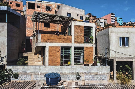 Fotografia colorida de uma casa em encosta urbana densamente ocupada. A construção tem estrutura aparente de concreto — pilares e vigas emoldurando os panos de parede — preenchida com alvenaria de tijolo cerâmico aparente. São dois pavimentos: embaixo, dois grandes caixilhos verdes de lâminas horizontais de vidro, entre panos de tijolo, com vasos de plantas na mureta à frente; em cima, um terraço com mureta de tijolo, onde está sentada uma pessoa, e, ao lado, outro caixilho verde igual. Sobre o terraço avança em balanço uma pérgola de ripas escuras apoiada em pilares metálicos finos. À esquerda, uma escada externa de concreto sobe até a casa, junto a vegetação e ao muro escuro do vizinho; à direita, uma casa vizinha de concreto sem pintura, com janela de caixilho claro e, embaixo, um vão aberto tomado por vegetação. Em primeiro plano, um muro de blocos e telhados de telhas onduladas, sobre os quais se apoia uma caixa-d’água azul. Ao fundo, o casario da comunidade sobe o morro — construções de tijolo sem acabamento, um prédio rosado e outro revestido de azul-claro — sob céu azul.