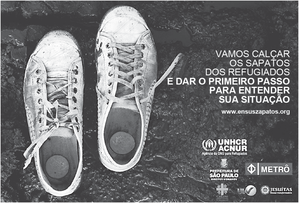 Cartaz em preto e branco com a fotografia, vista de cima, de um par de tênis brancos velhos, sujos e de cadarços desamarrados, sobre um chão escuro e molhado. À direita, em letras brancas: “VAMOS CALÇAR OS SAPATOS DOS REFUGIADOS E DAR O PRIMEIRO PASSO PARA ENTENDER SUA SITUAÇÃO”, seguido do endereço “www.ensuszapatos.org”. Abaixo, logotipos: UNHCR ACNUR – Agência da ONU para Refugiados, Prefeitura de São Paulo – Direitos Humanos, Metrô, Cáritas e Jesuítas, entre outros.