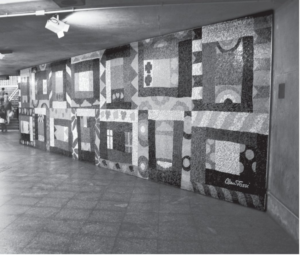 Fotografia em preto e branco de um corredor de estação de metrô com um grande painel de mosaico na parede: retângulos e quadrados de diferentes tonalidades e texturas dispostos como retalhos de uma colcha, com pequenos motivos (flores, corações, círculos, faixas listradas) dentro de alguns blocos. No canto inferior direito, a assinatura “Clau Tozzi”. À esquerda, uma escada com pessoas.