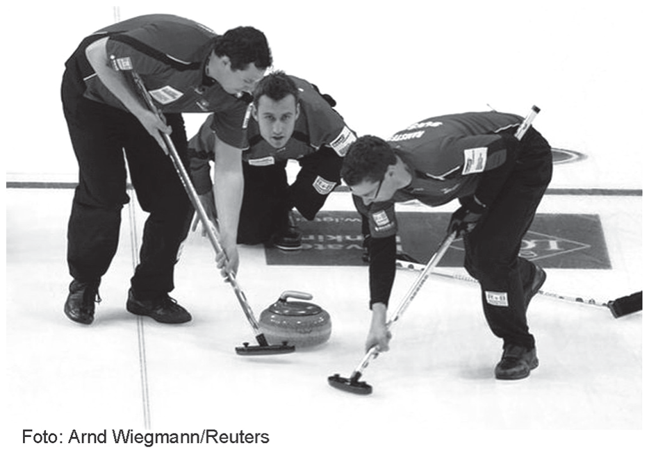 Fotografia em preto e branco de uma partida de curling: três atletas de uniforme escuro inclinados sobre a pista de gelo. Dois deles, à esquerda e à direita, esfregam vassouras no gelo logo à frente de uma pedra de granito com alça, que desliza entre eles; o terceiro, ao fundo, agachado, acompanha a jogada.